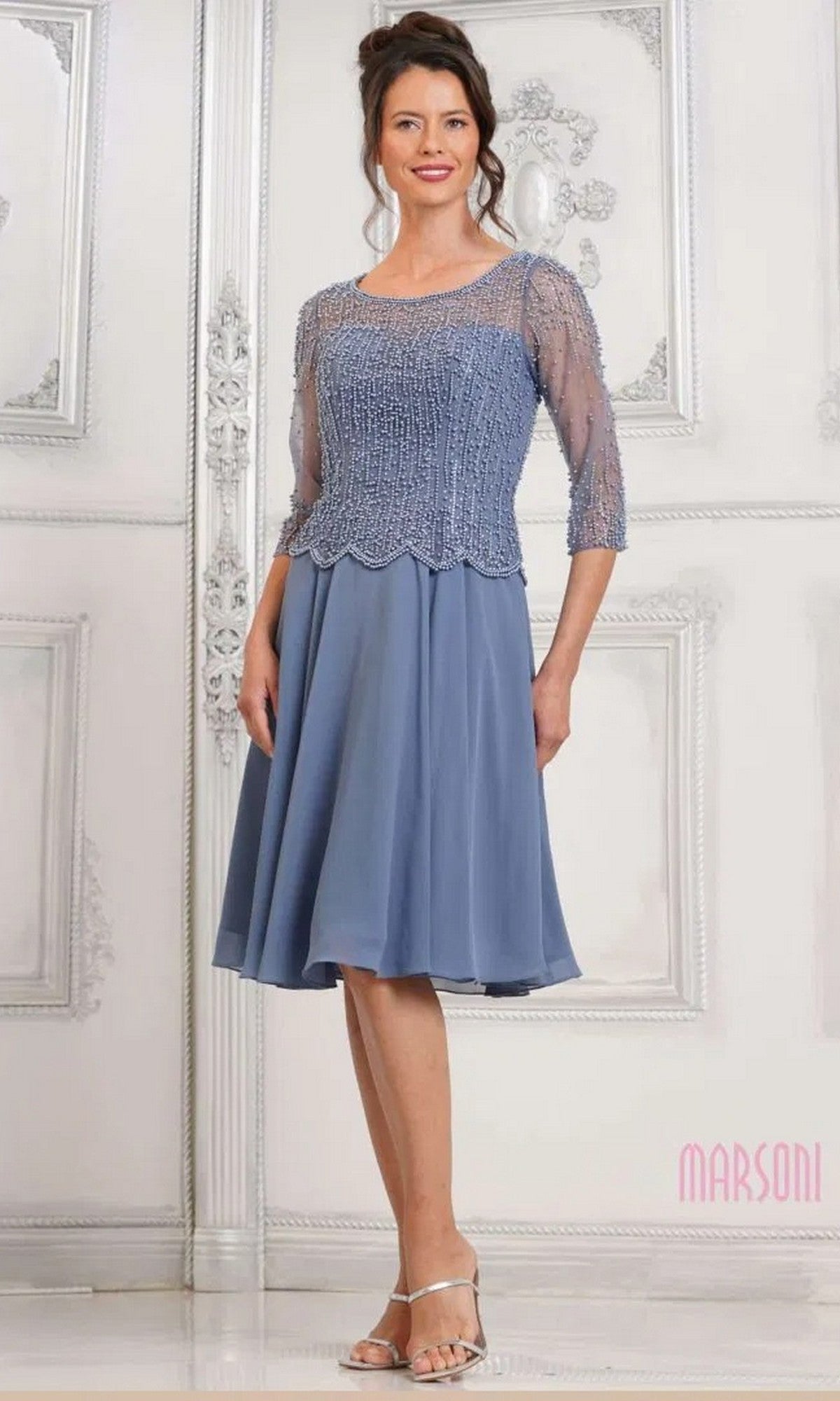 A woman stands in an ornate room with white paneled walls, wearing the Marsoni M312S knee-length semi-formal dress in slate blue with a chiffon A-line skirt and sheer, pearl-embellished half-sleeve top.